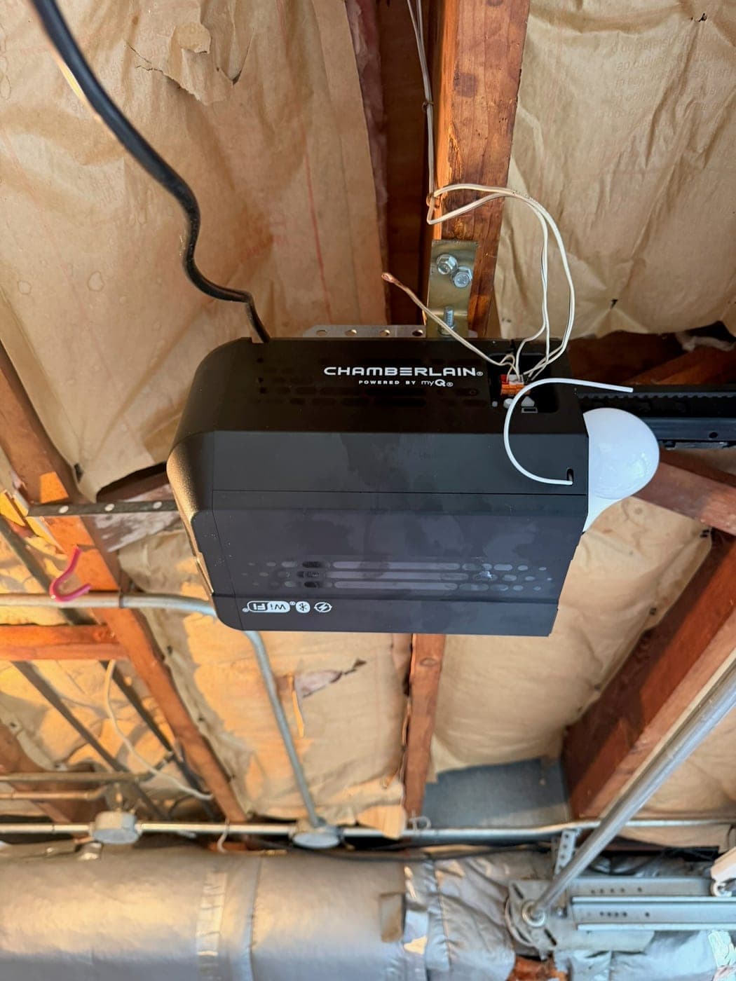 Chamberlain garage door opener mounted to exposed ceiling joists, with its light bulb and wiring